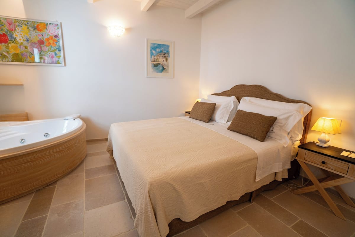 Een slaapkamer met een groot bed, een houten hoofdbord, een nachtkastje met een lamp, en een badkamer met een bad.