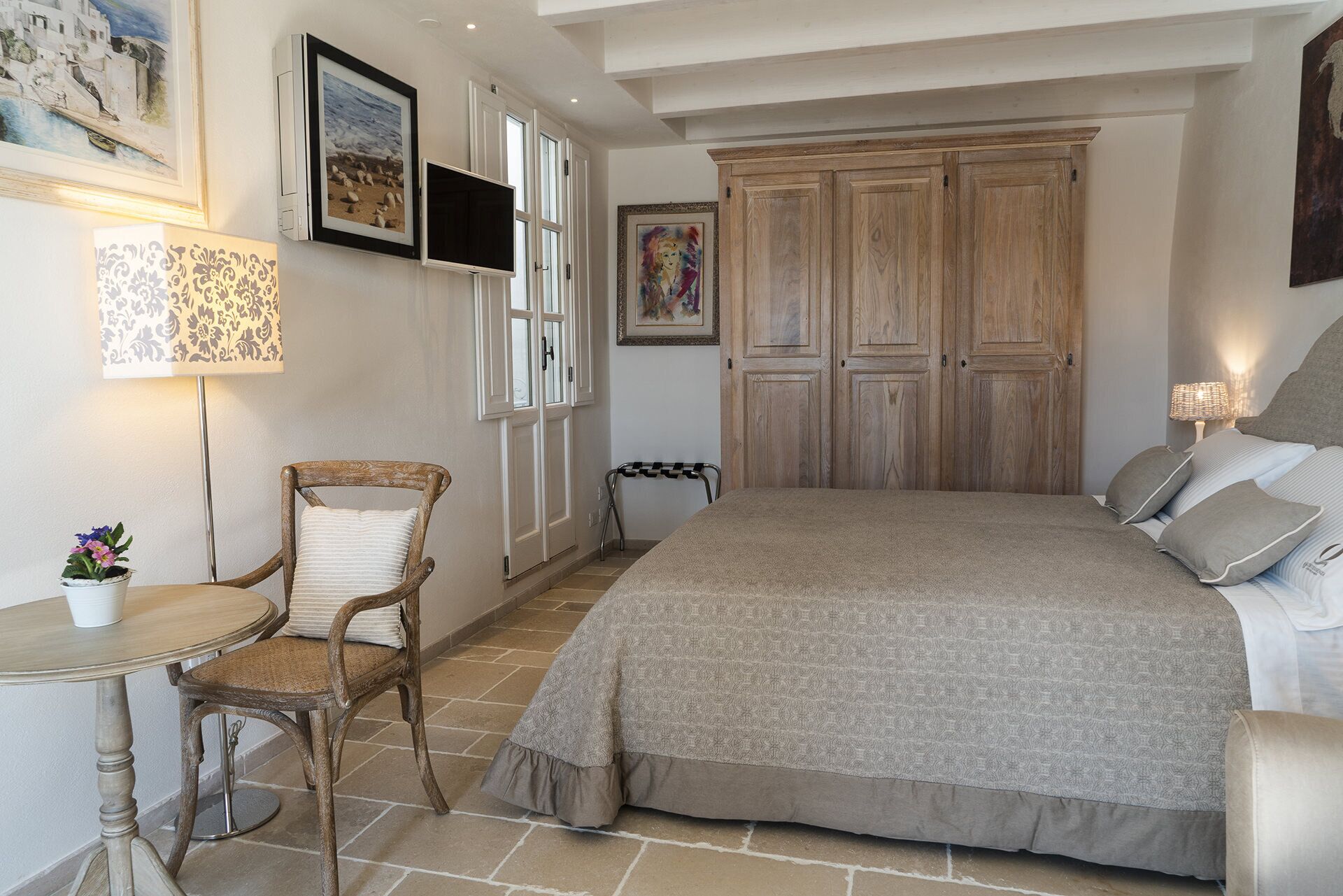 A bedroom with a large bed, a wooden wardrobe, a chair, a small table, and a lamp.