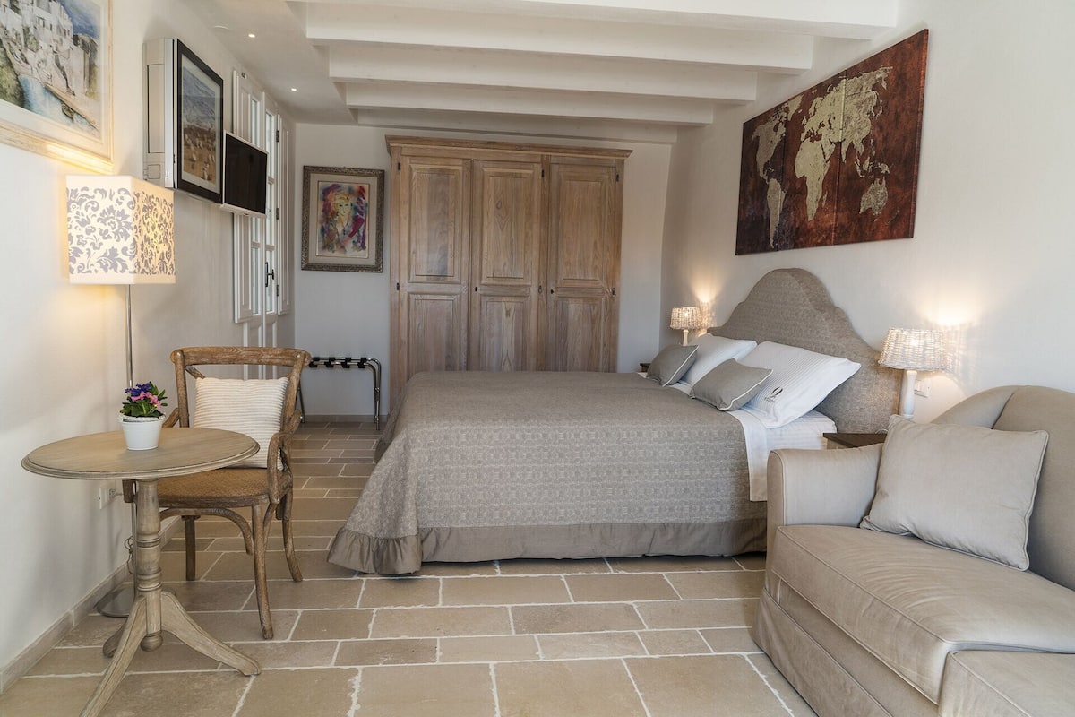 Een slaapkamer met een bed, een stoel, een klein tafeltje met een lamp en een bank.