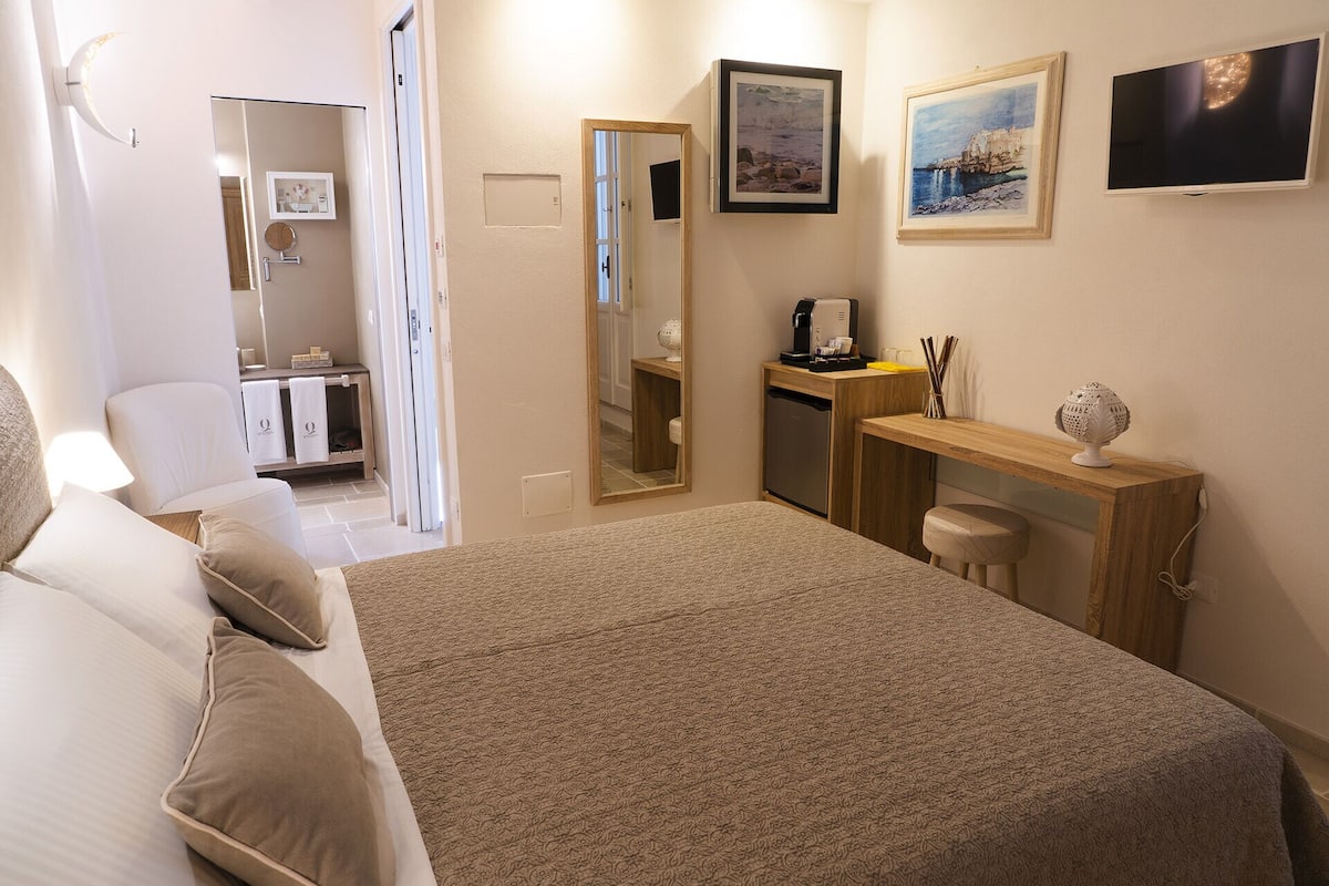 Een hotelkamer met een bed, een bureau, een stoel, een kleine koelkast, een koffiezetapparaat en kunstwerken aan de wanden.