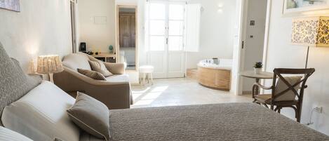 A bedroom with a large bed, a sitting area with a chair and table, and a view of the bathroom through an open door.