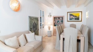 Een woonkamer met een beige bank, een eettafel met stoelen, een lamp, een boekenkast en een ingelijst schilderij aan de muur.