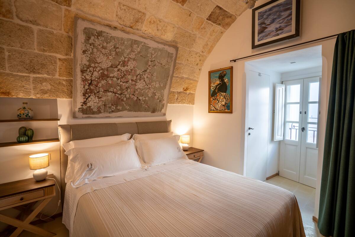 Een hotelkamer met een groot bed, een stenen muur, een schilderij en een deur naar een andere kamer.