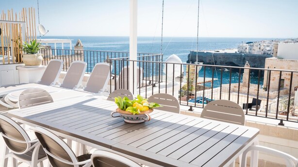 Een terras met een tafel, stoelen, een potplant en uitzicht op zee en gebouwen.