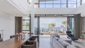 A modern living room with a large wooden dining table, comfortable seating, and a city view through floor-to-ceiling windows. - Penthouse 702 Kylemore B (Stellenbosch)