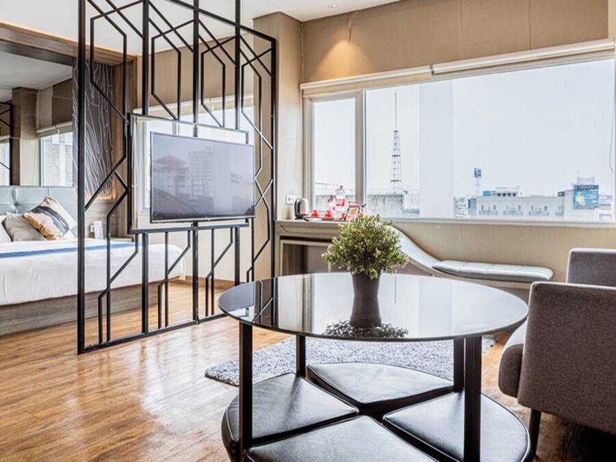 Una habitación de hotel moderna con cama, televisor, una mesa redonda con planta en maceta y un amplio ventanal con vistas a la ciudad.