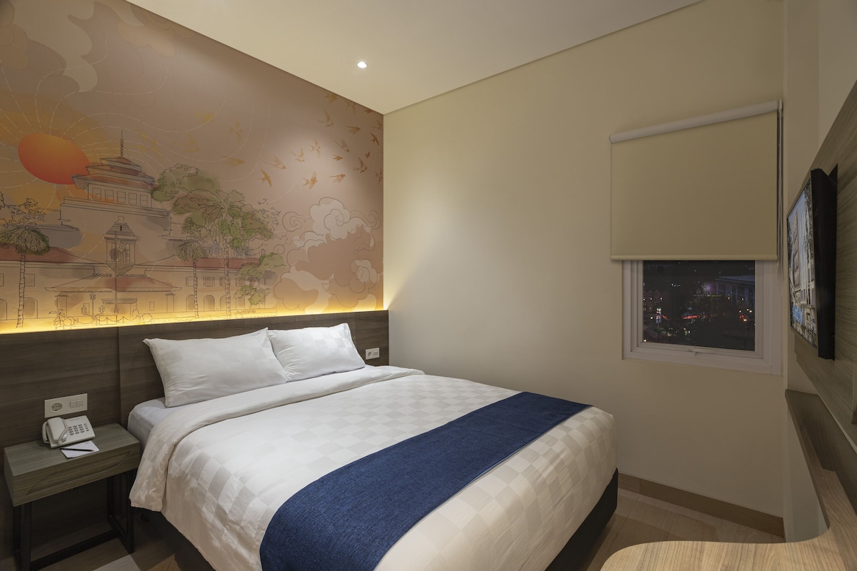 A hotel room with a large bed, a nightstand with a phone, a wall mural, a window with a view of the city, and a mounted TV.