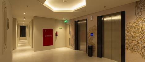 A hotel corridor with a modern design, featuring a red fire extinguisher, an elevator, and a wall with a decorative pattern.