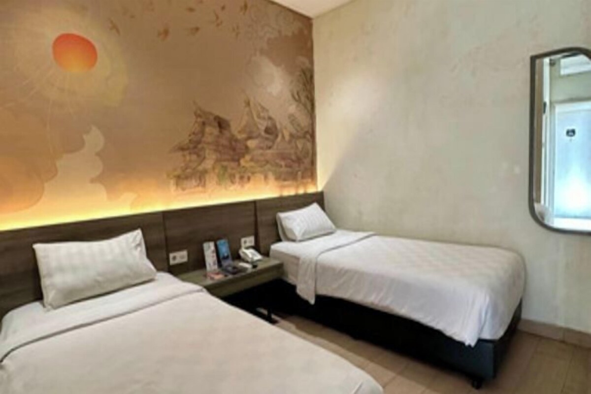A hotel room with two single beds, a nightstand with a phone and a magazine, a wall mural, and a window with a view.