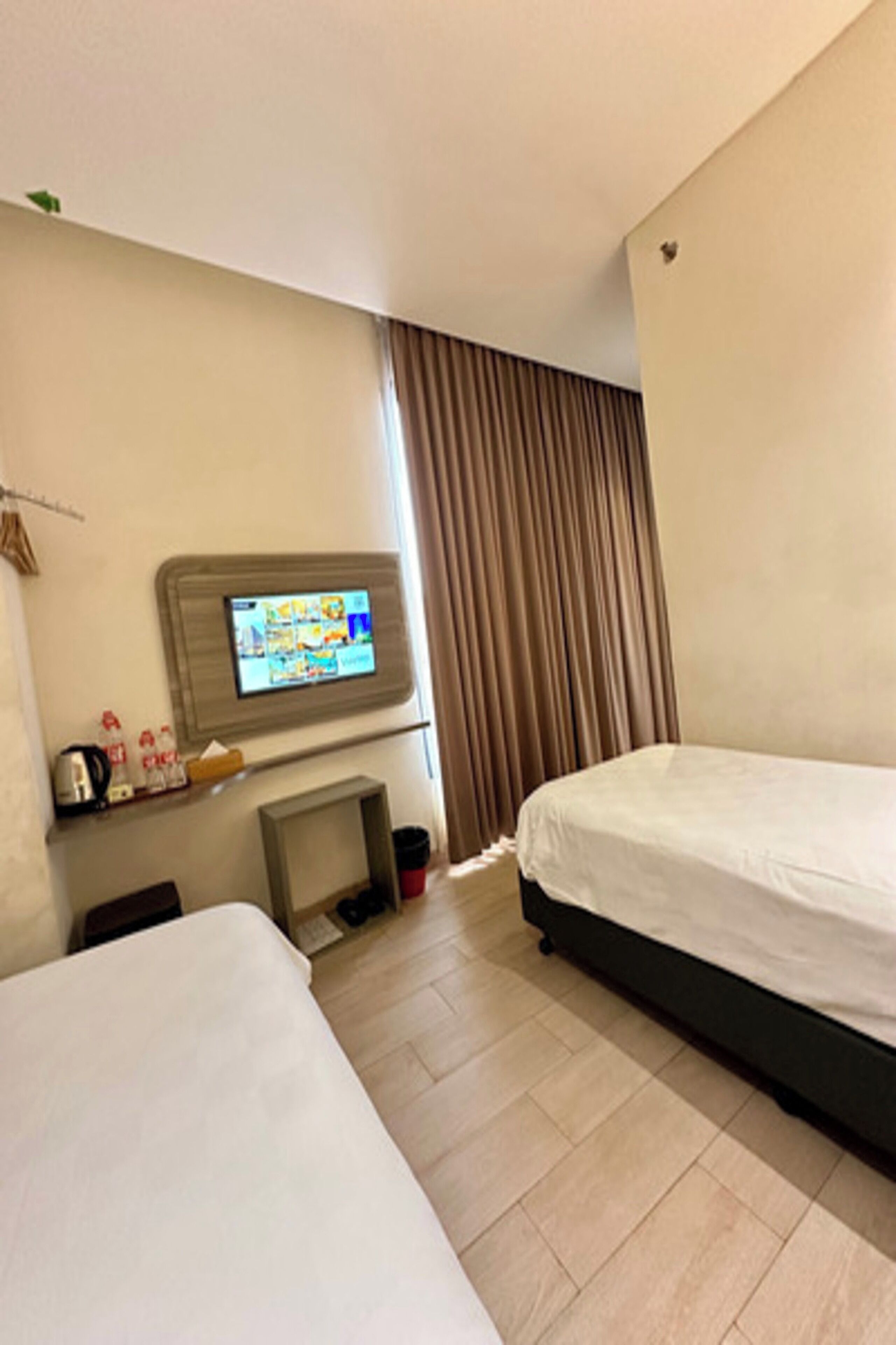 A hotel room with two beds, a television mounted on the wall, a small table with a kettle, and a window with curtains.