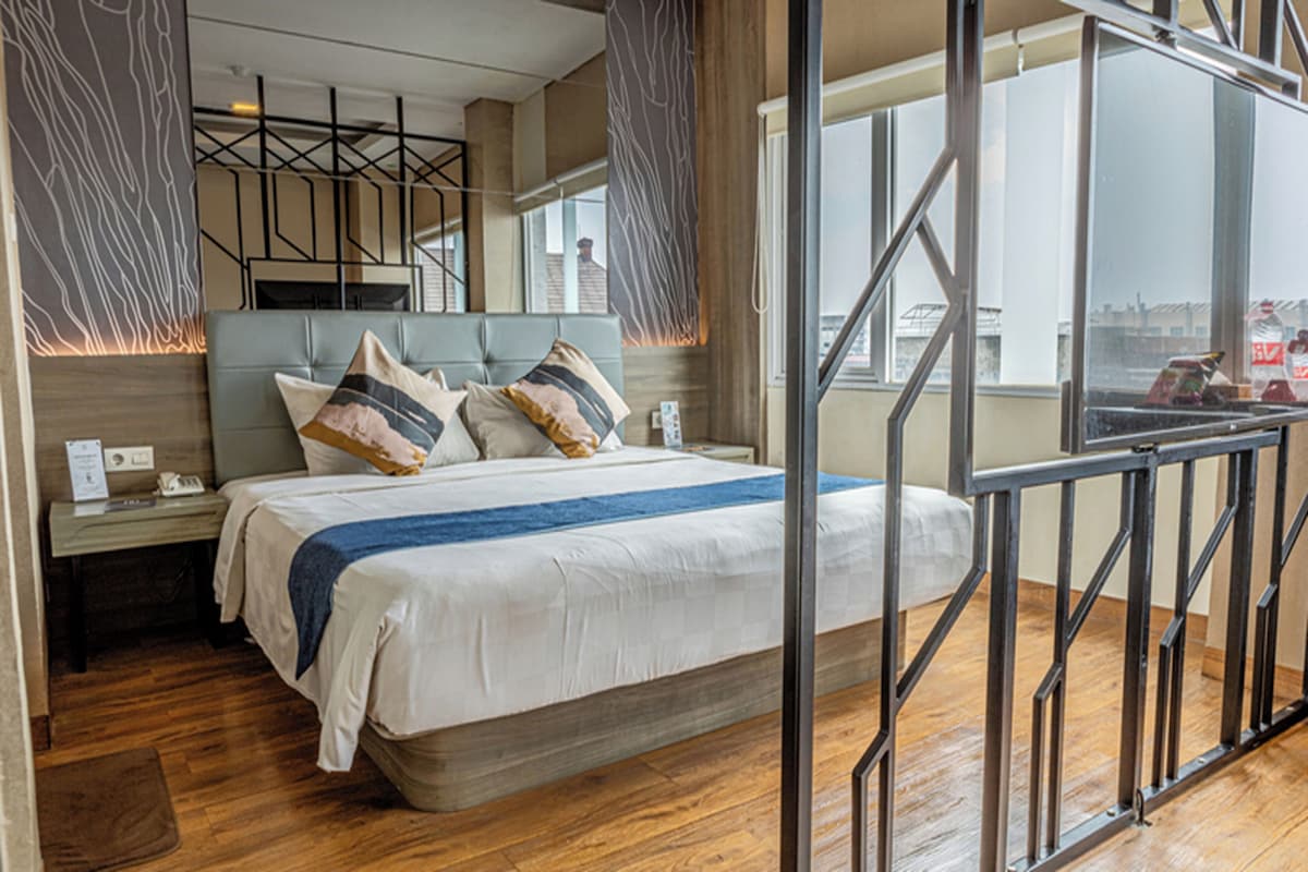 A modern hotel room with a large bed, a nightstand, a wooden floor, and a view of the city through large windows.