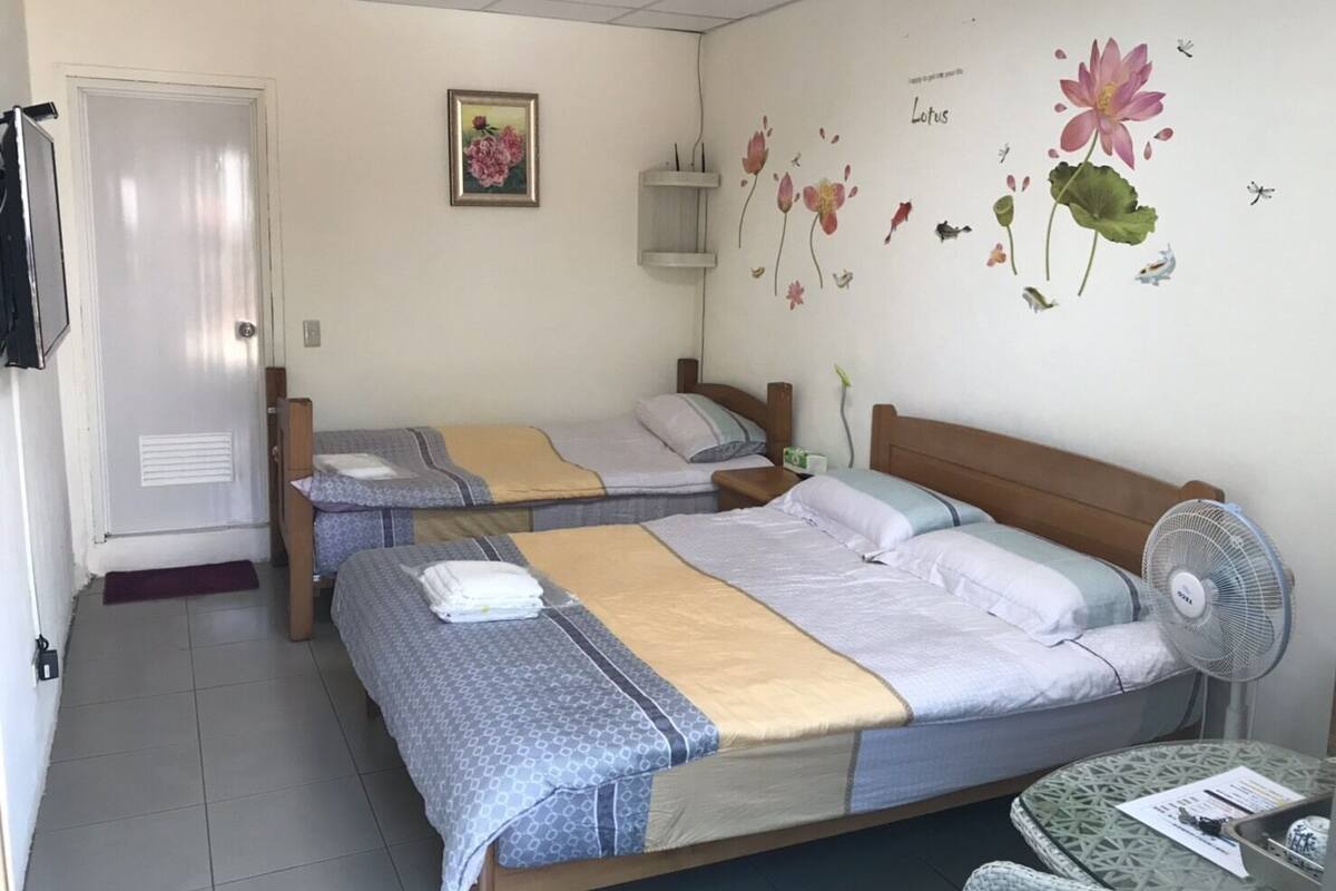 A room with two single beds, a small table, a fan, and a wall with floral decorations.