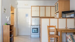 Eine kleine Küche mit Kühlschrank, Backofen und Holzschränken. Ein Stuhl und ein Tisch stehen dort, sowie ein Fernseher.