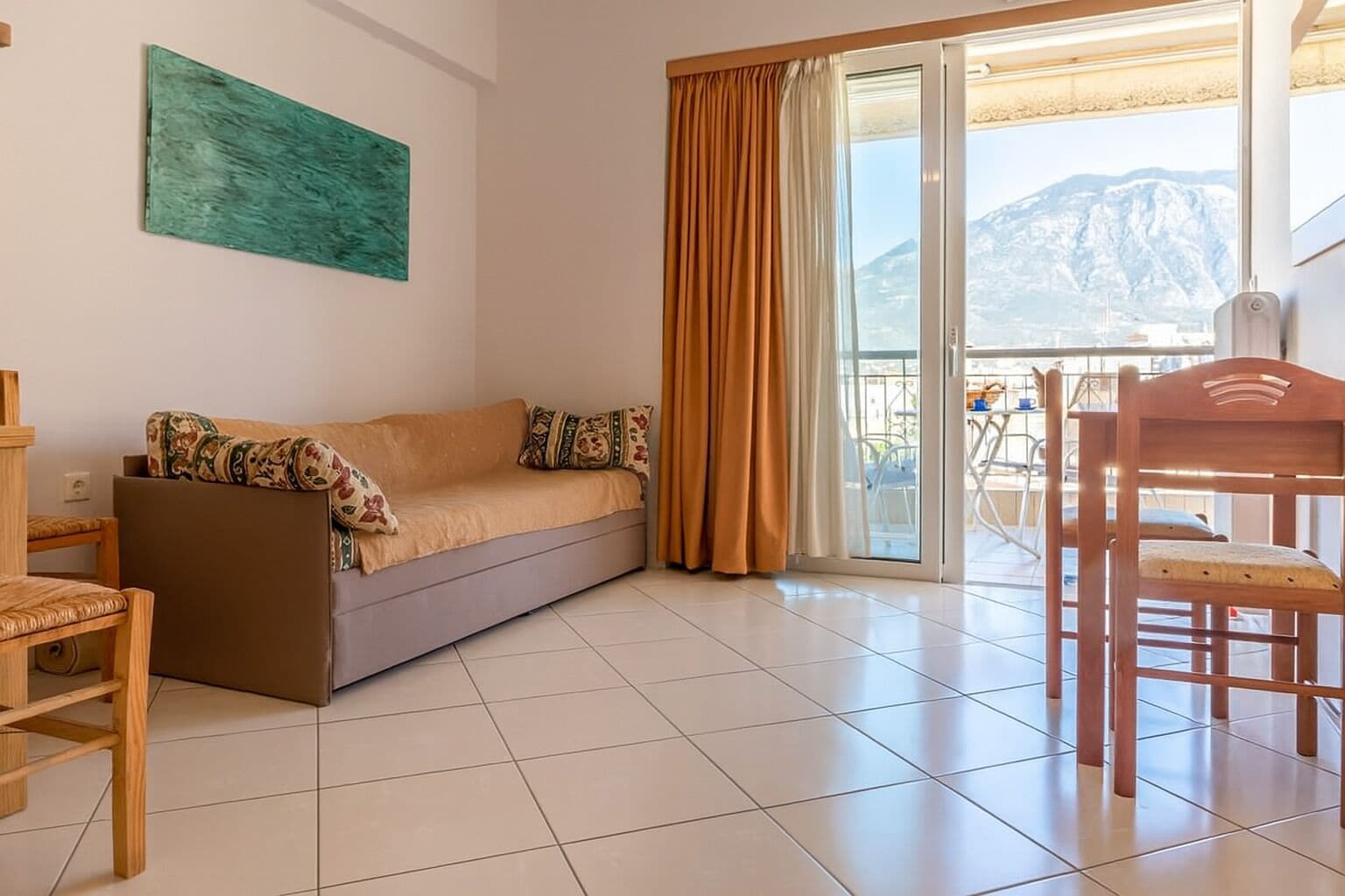 Una sala de estar con sofá, una silla de madera, una mesa, un ventanal con cortinas y vistas a la montaña.