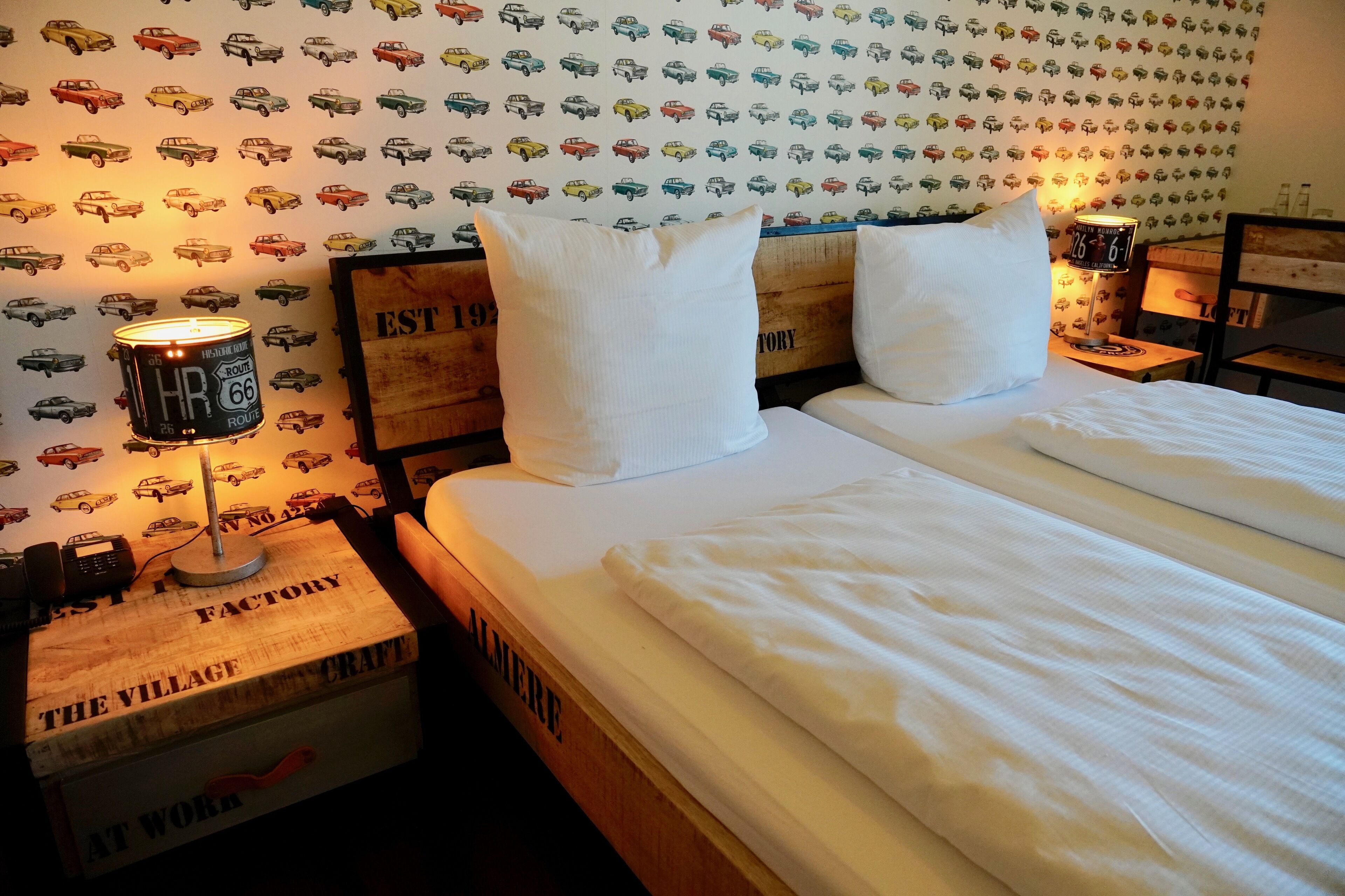 A neatly made bed with white linens, a headboard with a vintage design, and a wall decorated with a pattern of vintage cars.