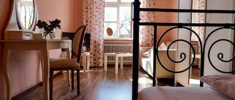 Un dormitori amb terra de fusta, un llit amb estructura de metall negre, un escriptori amb mirall, una cadira i una finestra amb cortines florals.