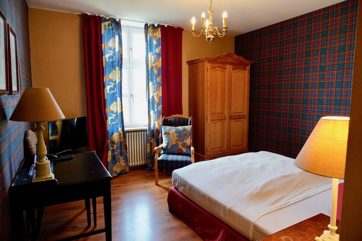 A hotel room with a bed, a desk with a lamp, a chair, a wardrobe, and a television.