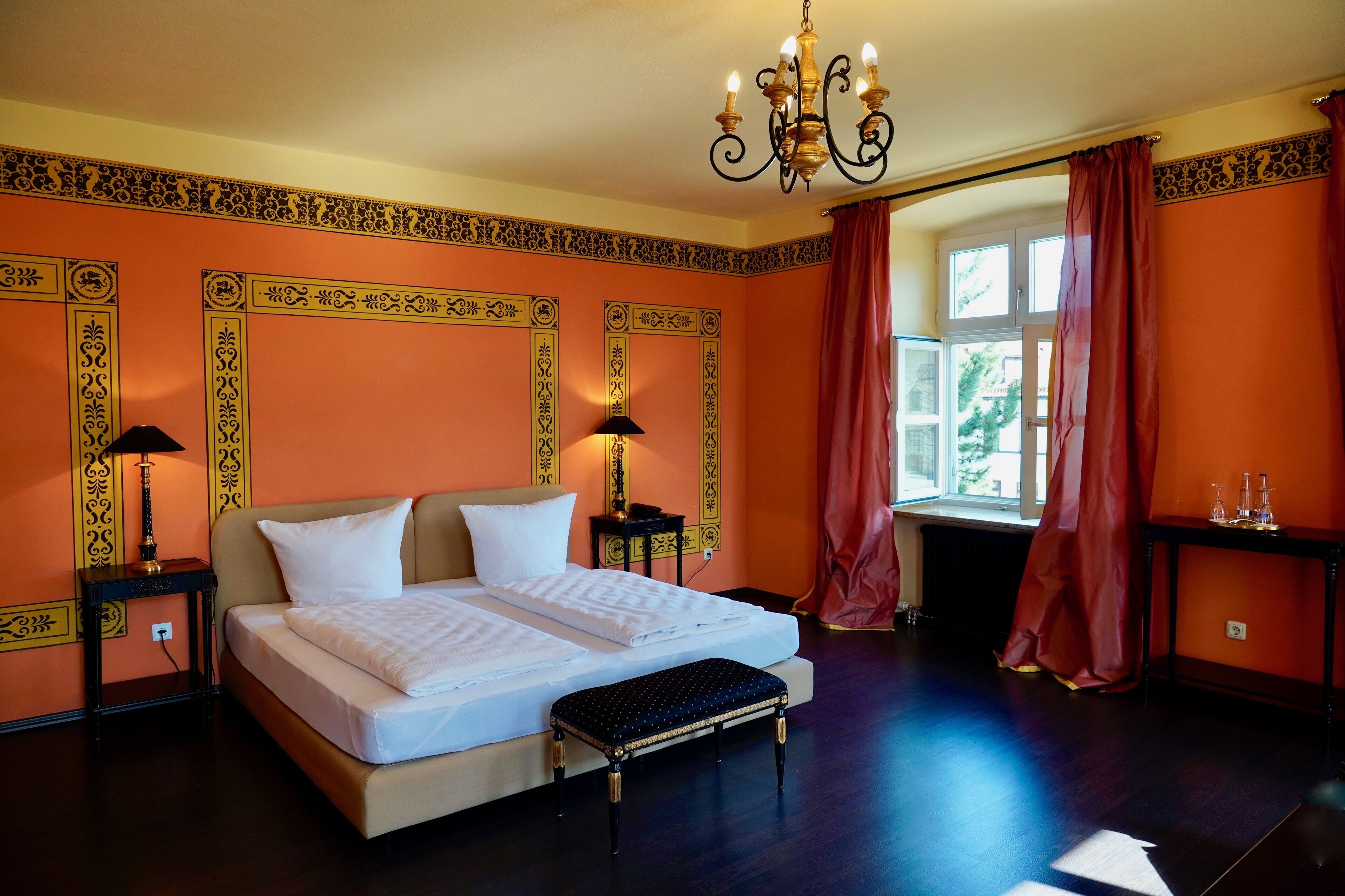 A hotel room with a large bed, a bench, a chandelier, and a window with curtains.