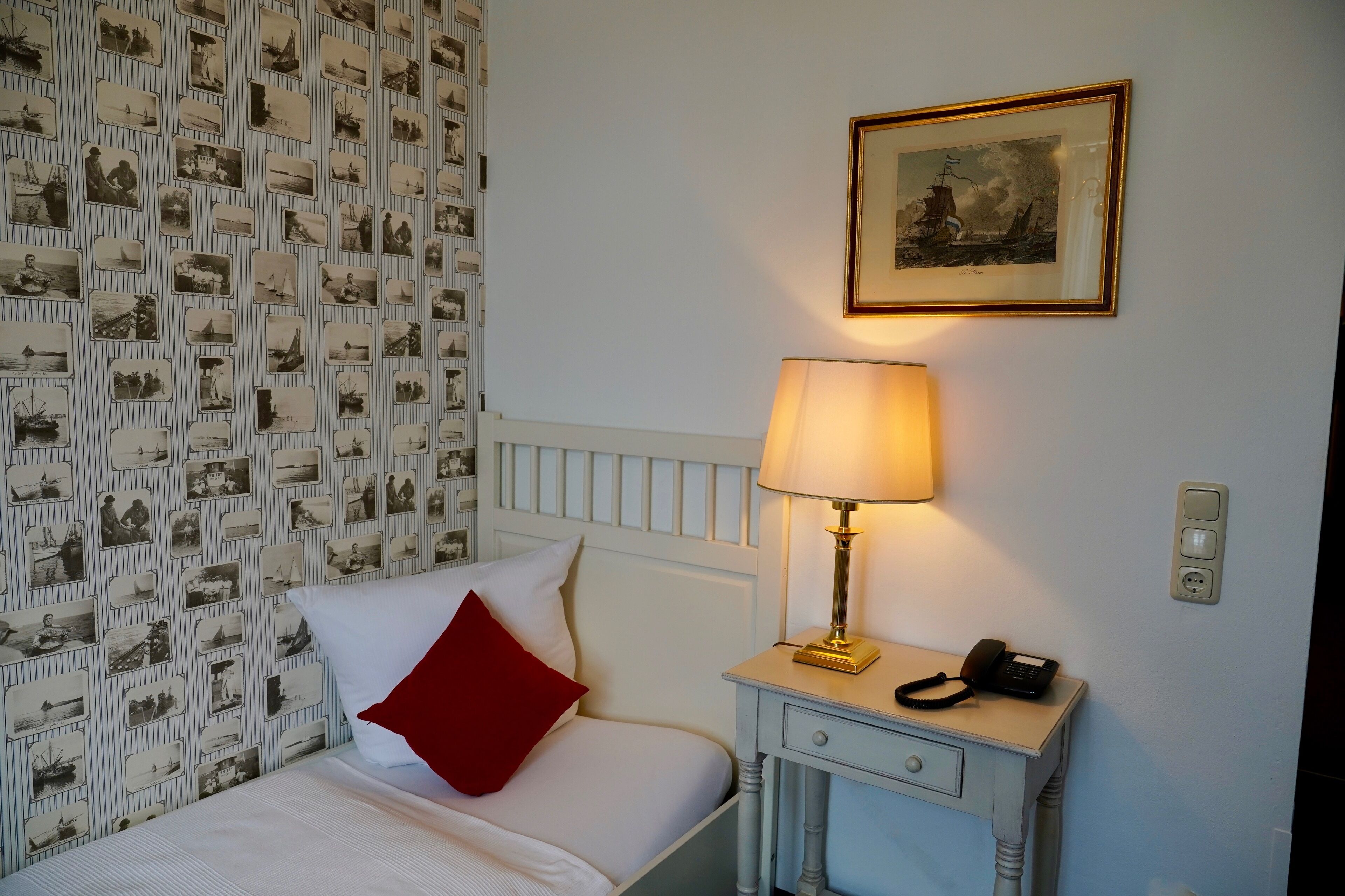 Una cama bien hecha con almohada blanca y roja, una mesita de noche con lámpara y teléfono, y una pared con un cuadro enmarcado.