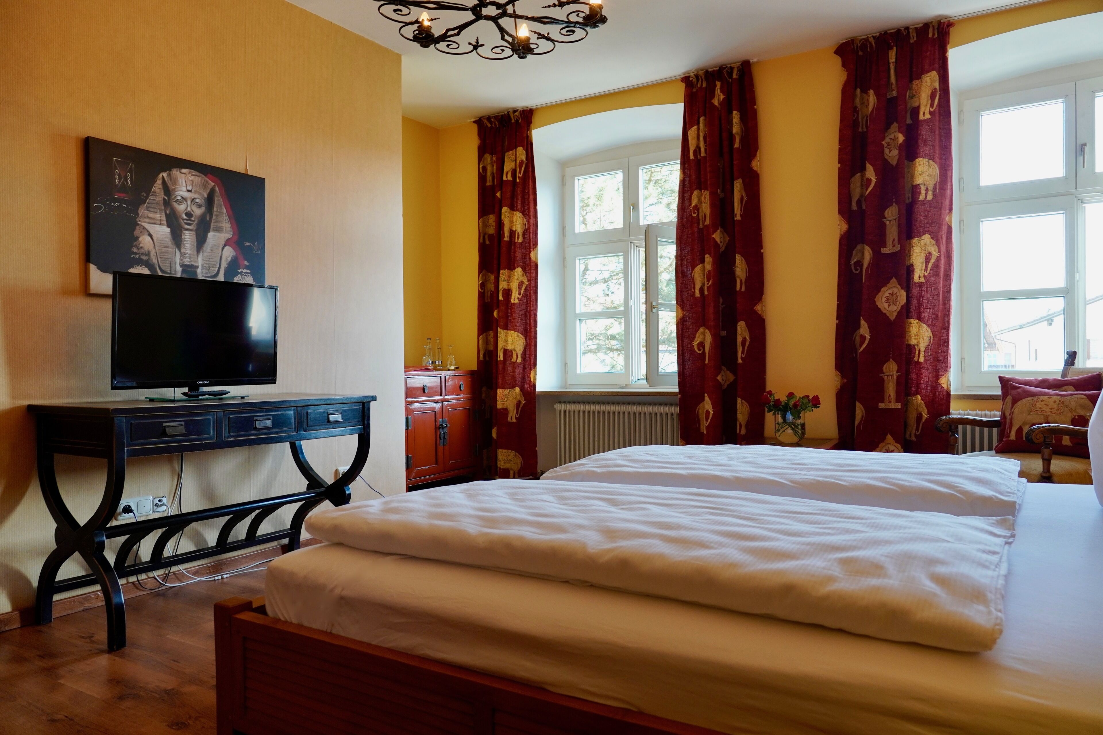 Quarto com cama, escrivaninha com televisão, janela com cortinas e radiador.
