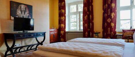 A bedroom with a bed, a desk with a television, a window with curtains, and a radiator.