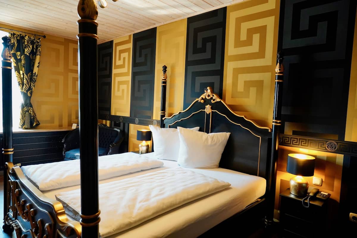 A four-poster bed with white linens, a nightstand with a lamp, and a wall with a geometric pattern.