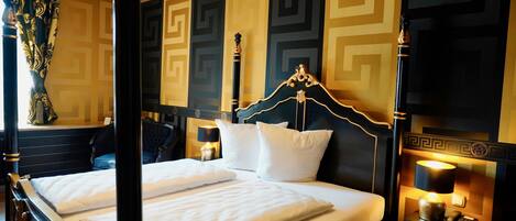 A four-poster bed with white linens, a nightstand with a lamp, and a wall with a geometric pattern.