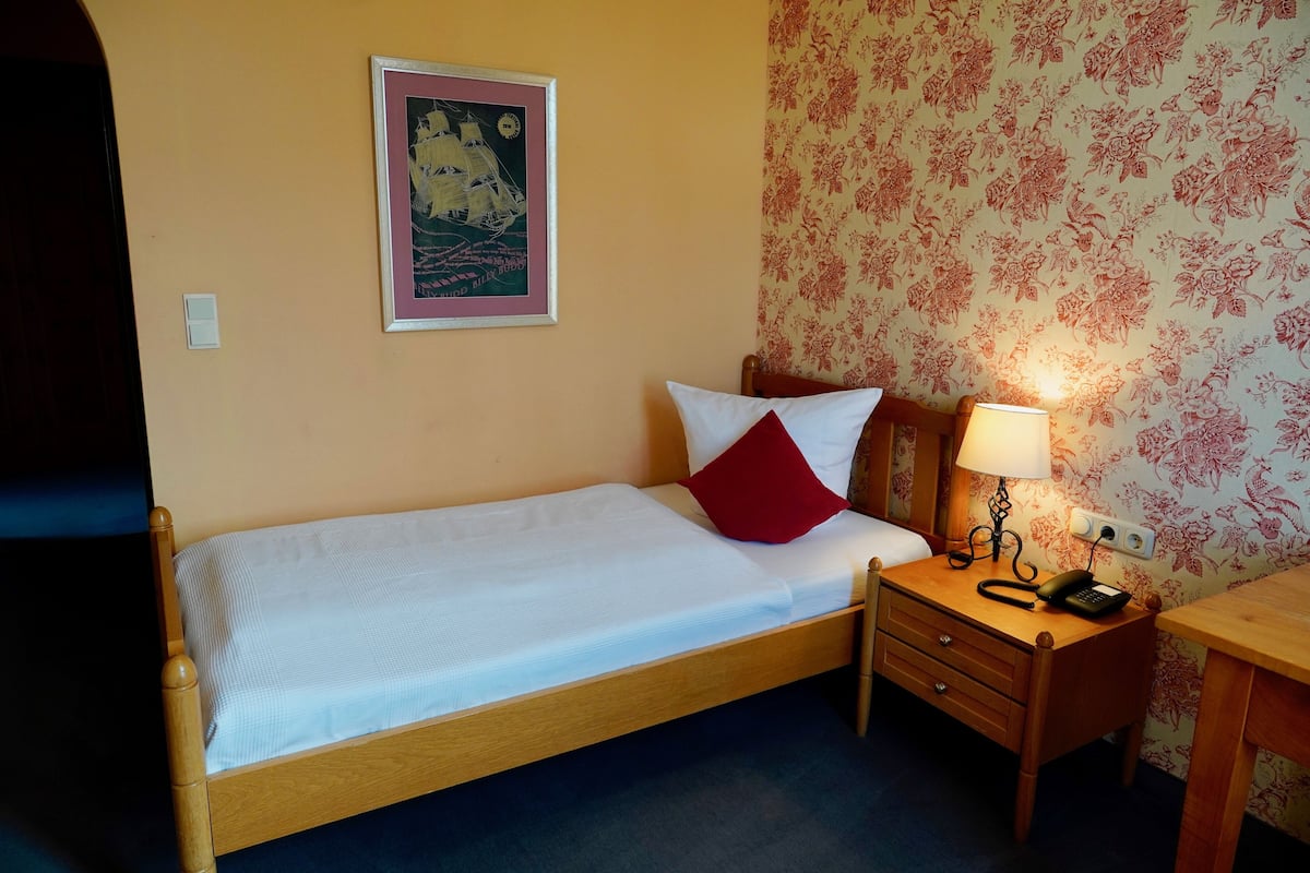 A single bed with a white and red pillow, a bedside table with a lamp, and a wall with floral wallpaper.