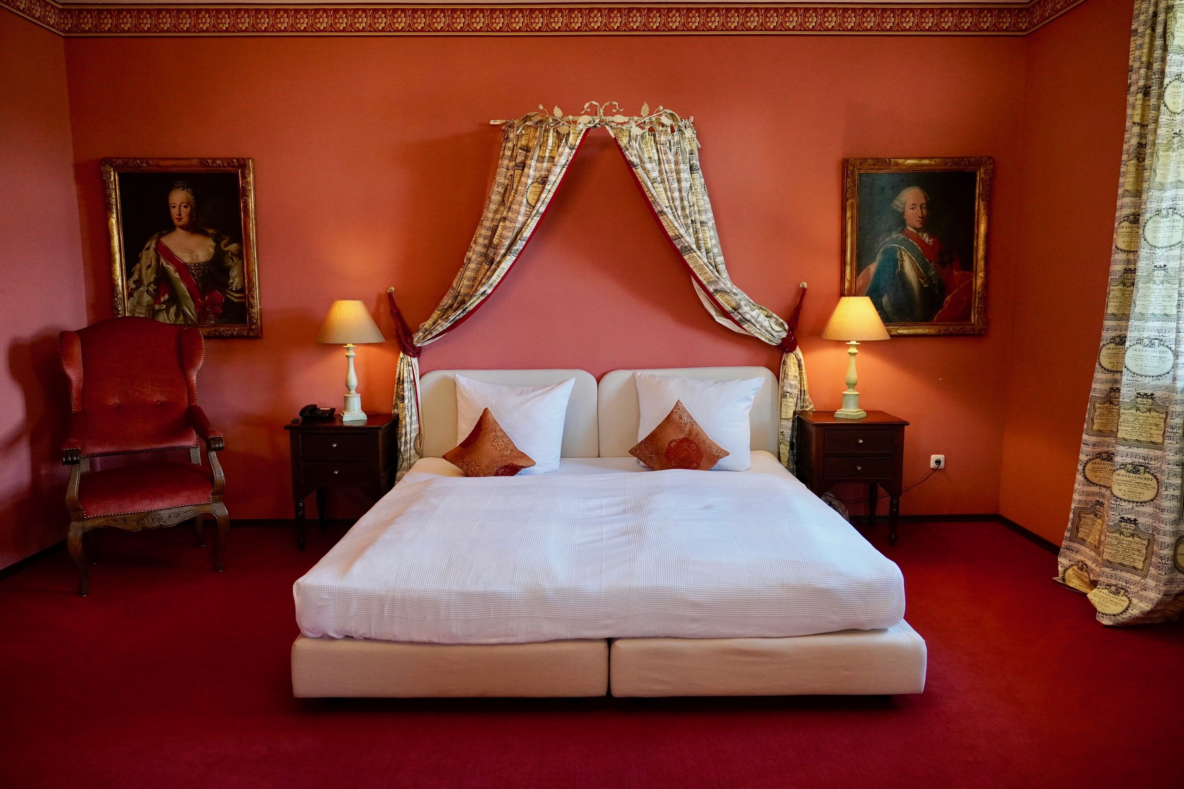 Habitación de hotel con una cama grande, dos mesitas de noche, un sillón rojo y dos cuadros en las paredes.