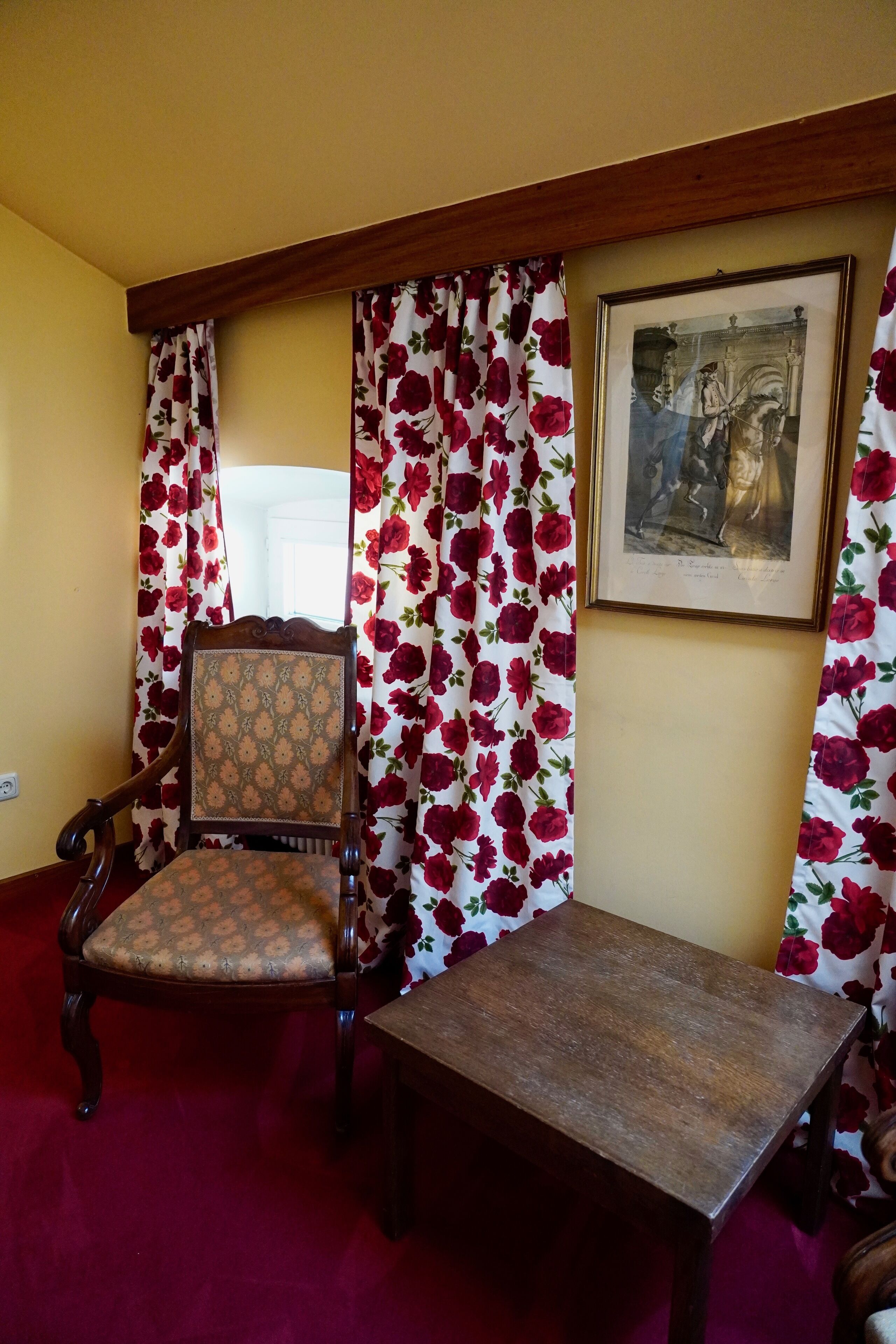 A room with a wooden chair and a table, floral curtains, and a framed picture on the wall.