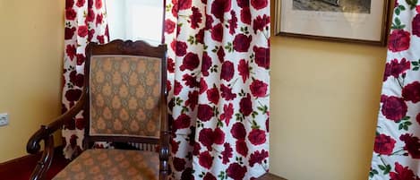 A room with a wooden chair and a table, floral curtains, and a framed picture on the wall.