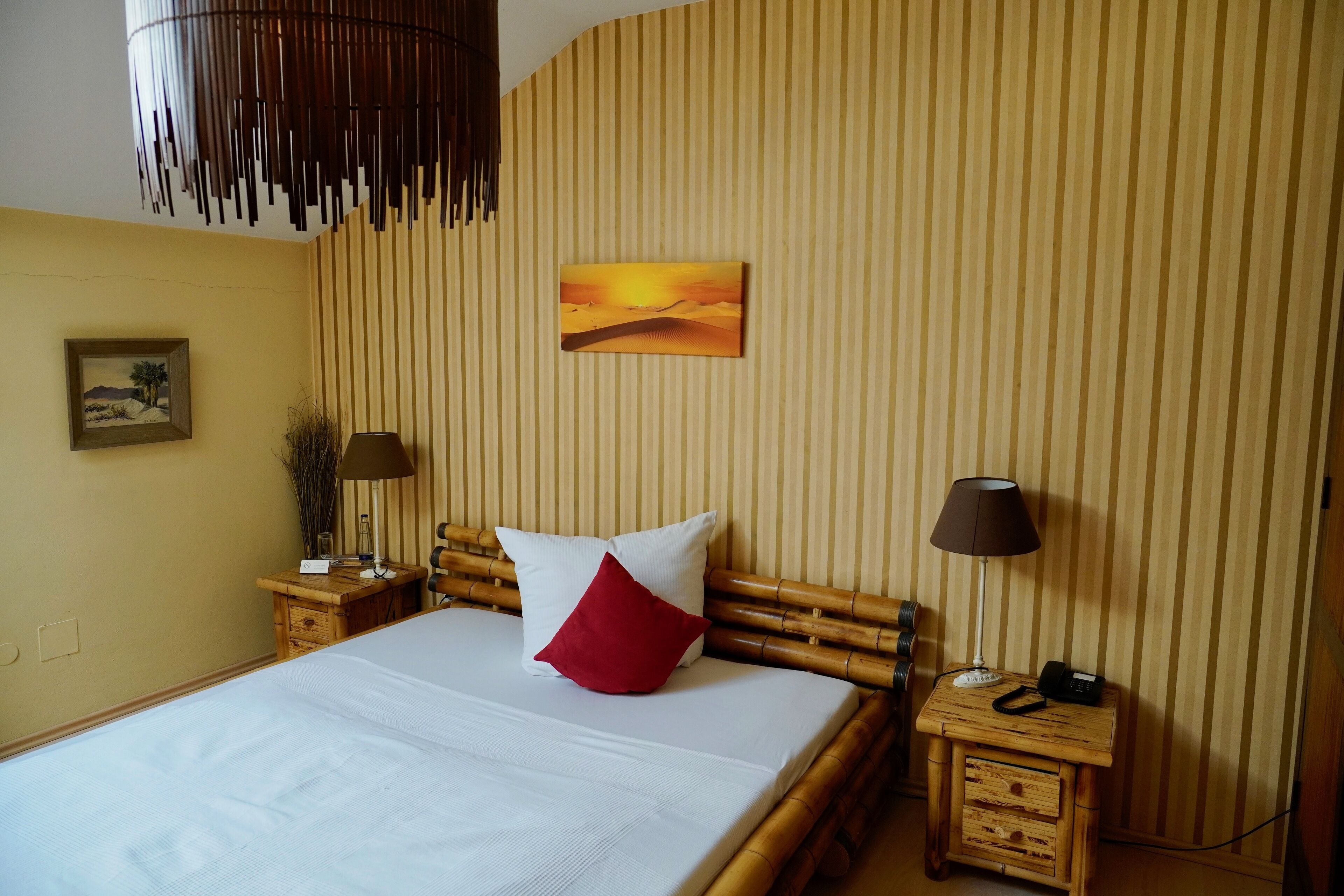 Uma cama bem arrumada com almofadas brancas e vermelhas, cabeceira de madeira e uma pequena mesa de cabeceira com uma luminária e um telefone.