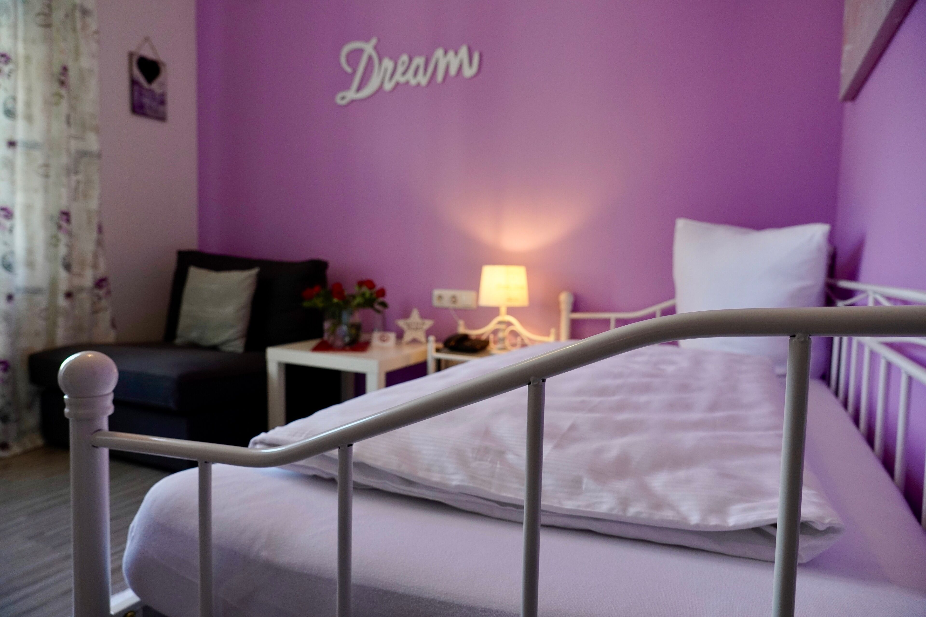 Une chambre avec un mur violet, un lit blanc et une petite table de chevet avec une lampe.