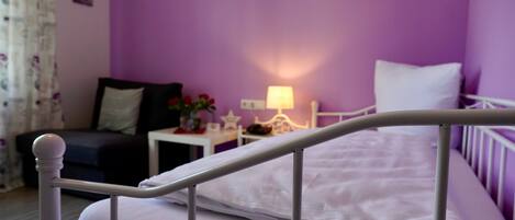 Un dormitorio con una pared morada, una cama blanca y una mesita de noche pequeña con una lámpara.