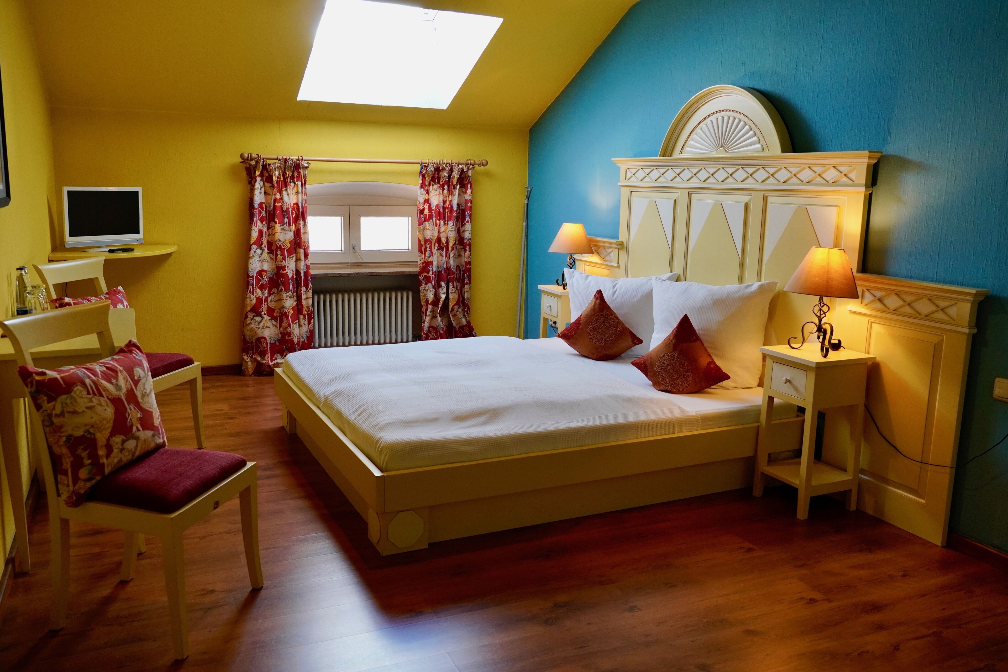 Une chambre à coucher comprenant un lit, des tables de chevet, une chaise, un bureau, une télévision et une fenêtre avec des rideaux.
