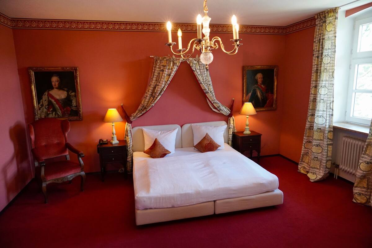 A bedroom with a large bed, two bedside tables with lamps, a red armchair, and two paintings on the walls.