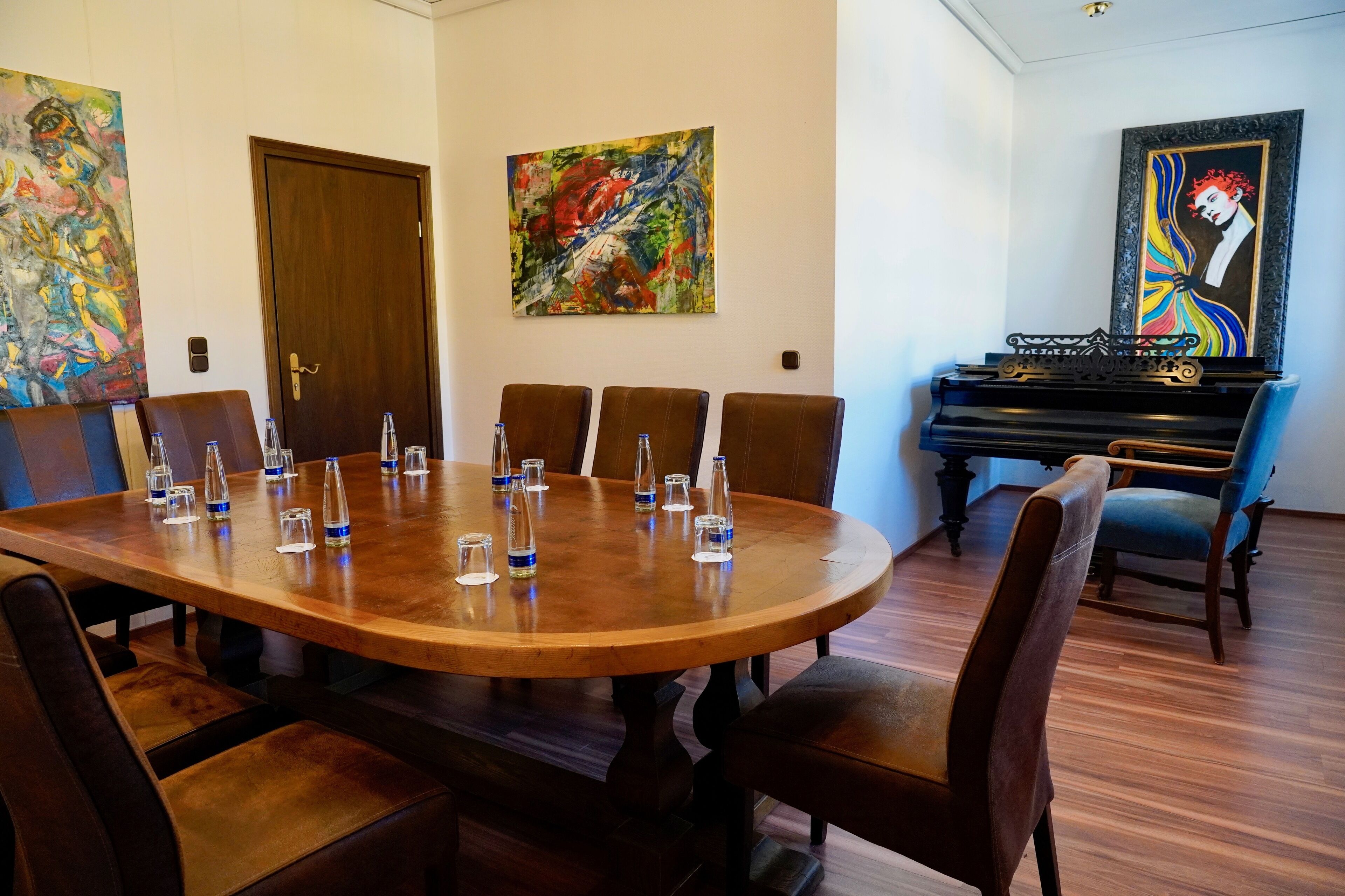 Een vergaderruimte met een houten tafel, zes stoelen en waterglazen. Twee schilderijen hangen aan de muur en er staat een piano op de achtergrond.