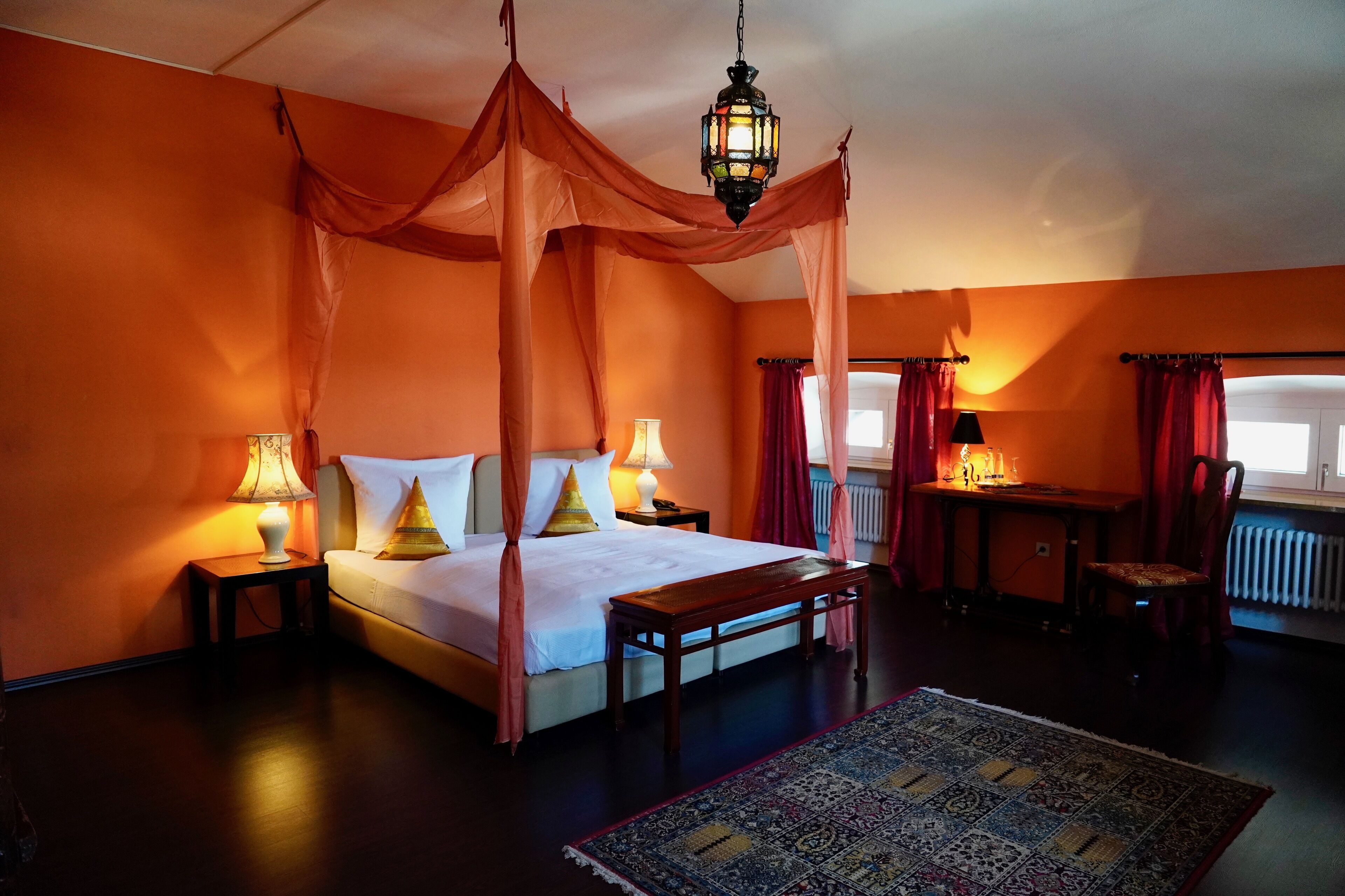 Ein Schlafzimmer mit einem Himmelbett, zwei Nachttischen mit Lampen, einer Holzbank, einem gemusterten Teppich und Blick durch ein Fenster mit Vorhängen.