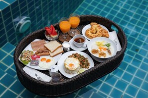 Un plateau de petit-déjeuner contenant divers plats, notamment des œufs, du riz, des toasts et des fruits, placé sur une table au bord de la piscine.