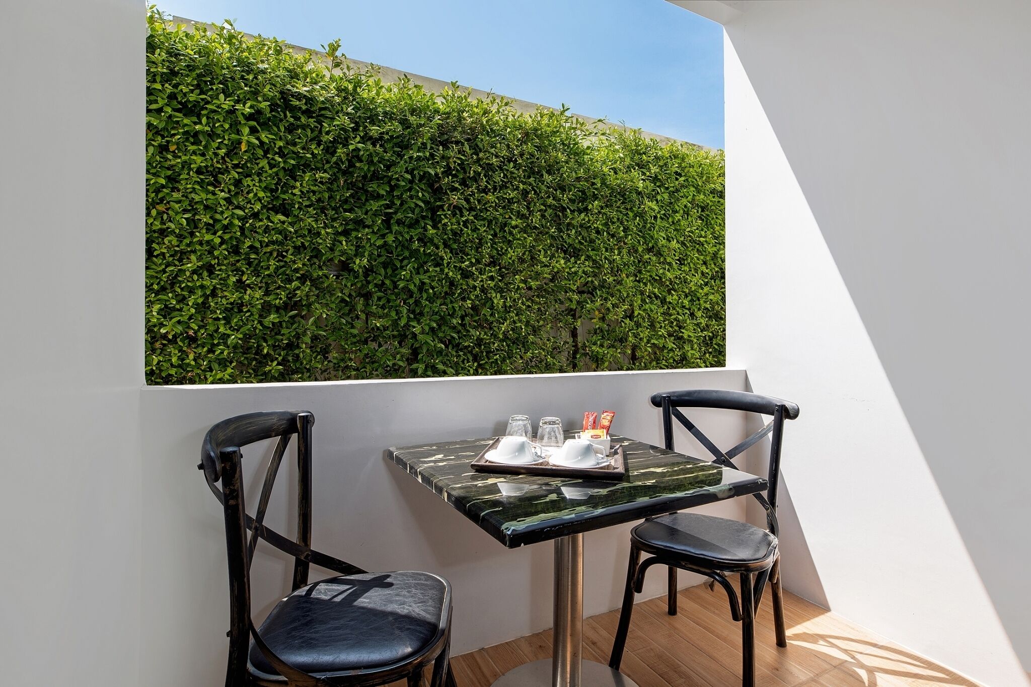 Un balcón con una mesa y dos sillas, un muro de seto verde y un cielo despejado.