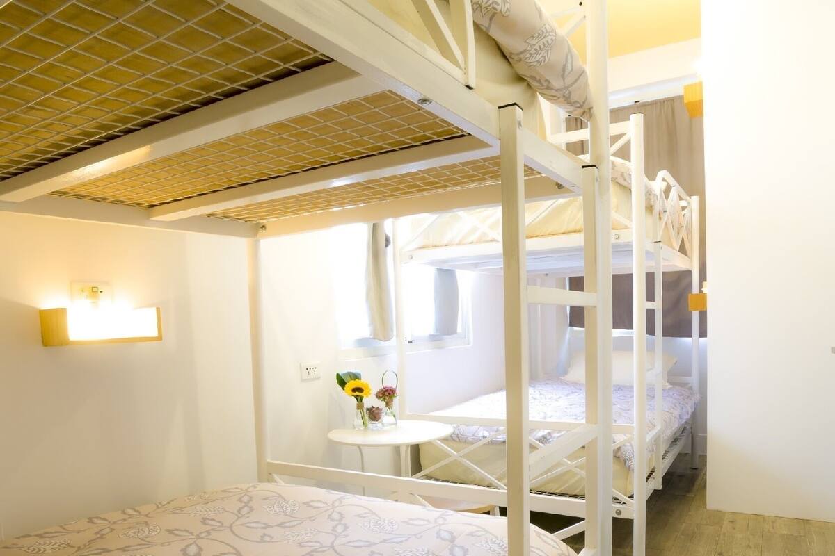 Une chambre avec un lit superposé, une couvre-lit à motifs floraux, une petite table avec un vase de fleurs et une fenêtre avec des rideaux.