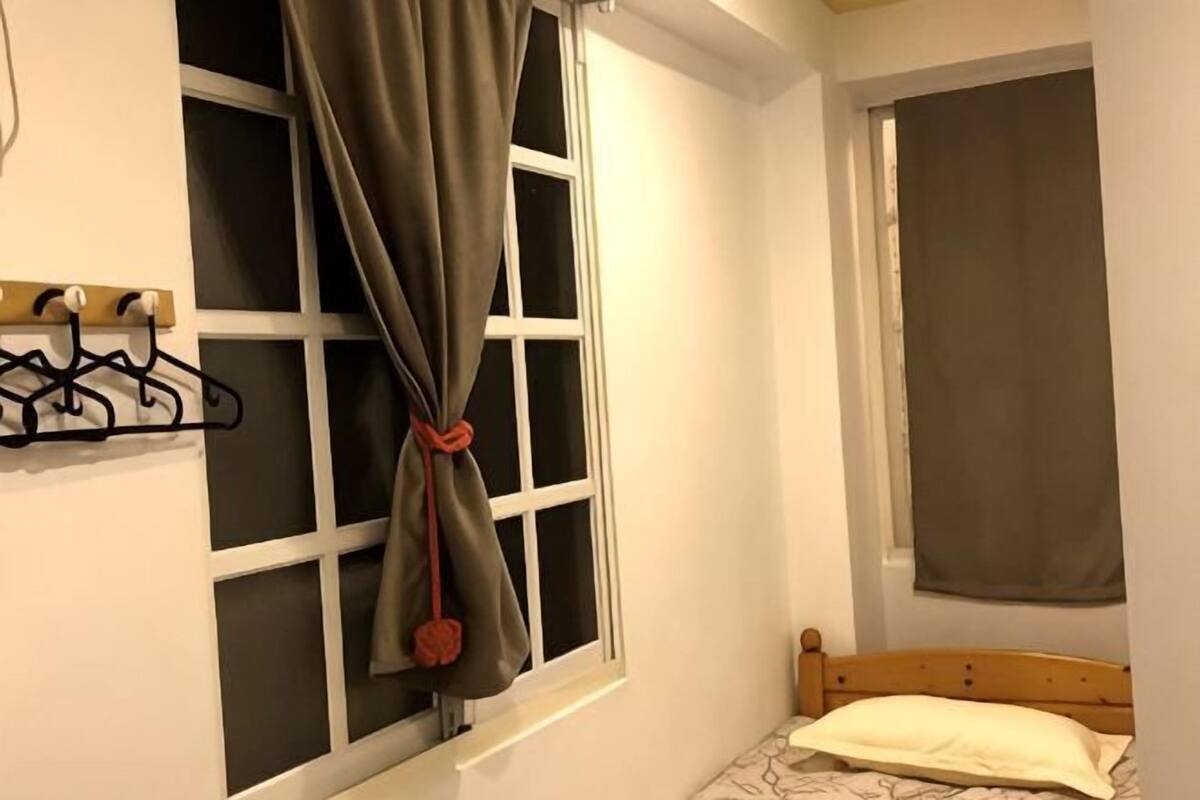 Une chambre à coucher avec un lit, une fenêtre avec un rideau retenu par un cordon, une tête de lit en bois et un porte-manteau fixé au mur.