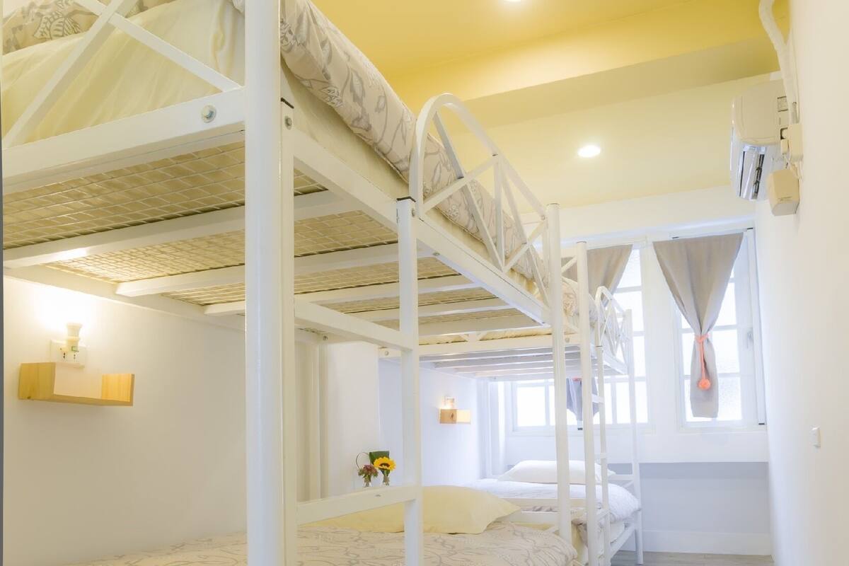 Une chambre avec un lit superposé, un plancher en bois, une petite étagère avec une lampe et une fenêtre avec des rideaux.