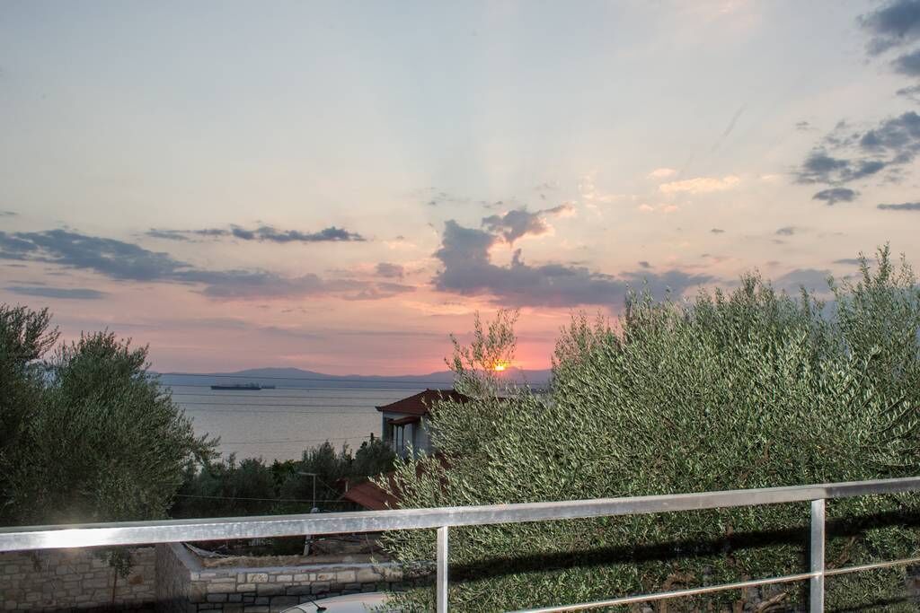 A coastal view with a sunset, a body of water, and a building partially obscured by foliage.