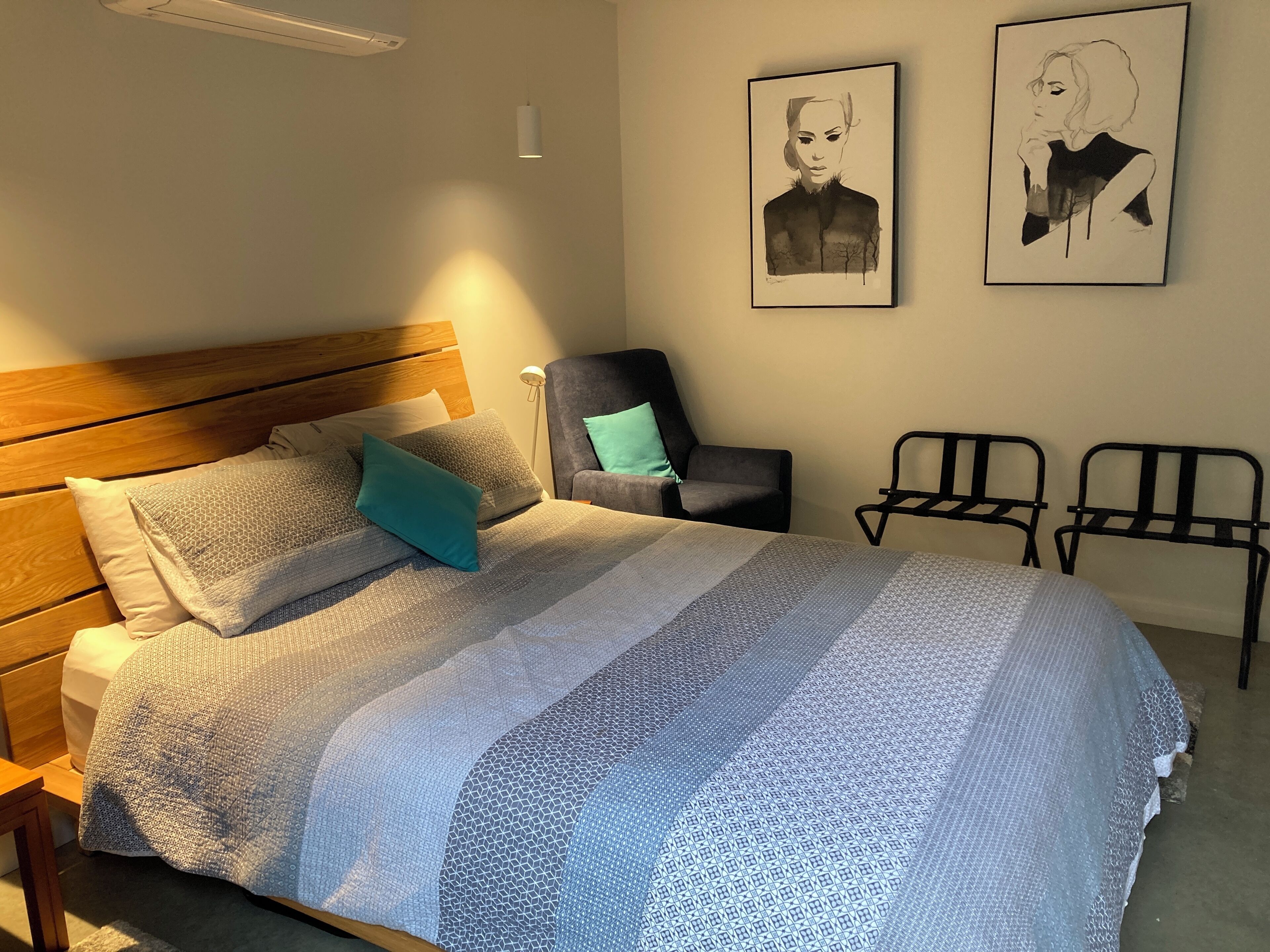 Een opgemaakt bed met een geborduurd deken, een houten hoofdbord en een teal-kleurig kussen. Twee ingelijste kunstwerken hangen aan de muur, een stoel en een kleine tafel staan naast het bed.