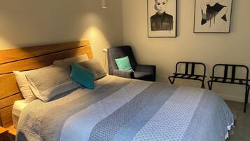 Een opgemaakt bed met een geborduurd deken, een houten hoofdbord en een teal-kleurig kussen. Twee ingelijste kunstwerken hangen aan de muur, een stoel en een kleine tafel staan naast het bed.