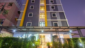 Un hôtel à plusieurs étages au design moderne, caractérisé par une façade centrale aux couleurs jaune et gris, de nombreuses fenêtres et un espace d’entrée abrité agrémenté de plantes en pot.