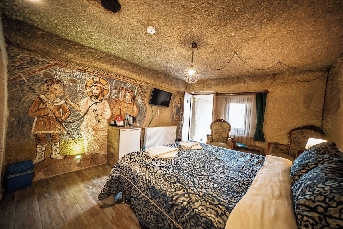 A room with a large mural, a bed with patterned bedding, a television, and a chair.