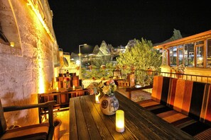 An outdoor seating area with wooden tables and chairs, a vase with flowers, and a view of a mountain range at night. - Adelya Cave Hotel (Nevsehir)