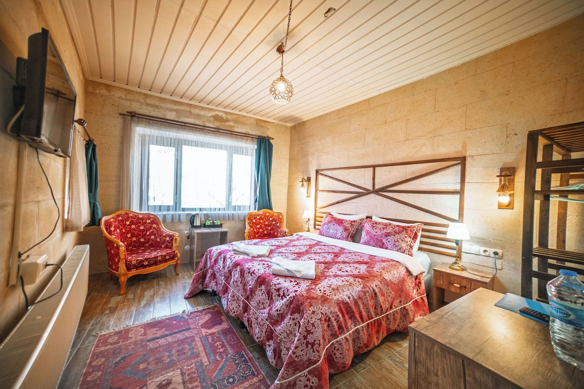A bedroom with a wooden ceiling, a large bed with a patterned bedspread, a red armchair, a wooden nightstand with a lamp, and a window with curtains.